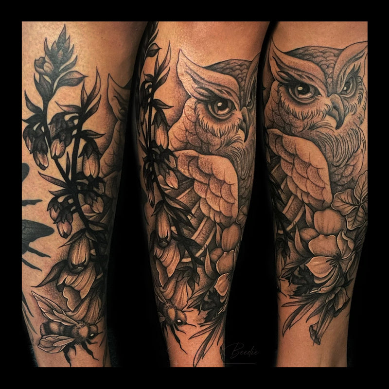 Detailed black and grey lower leg calf tattoo of a illustrative, realistic owl surrounded by flowers, leaves, and a bee in a botanical style