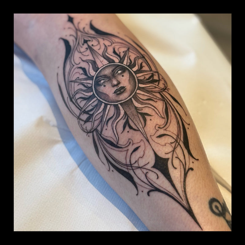 Black and grey calf, leg tattoo of a stylized sun with a feminine face, surrounded by flowing ornamental symmetrical fine linework